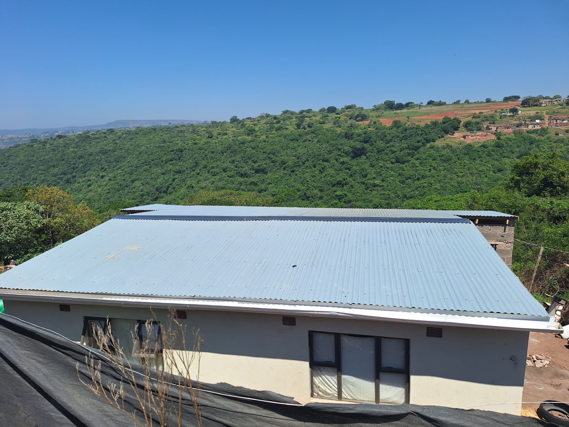 A new corrugated metal roof covering Baba Buthelezi’s home with the green hills of Ntshongweni behind it