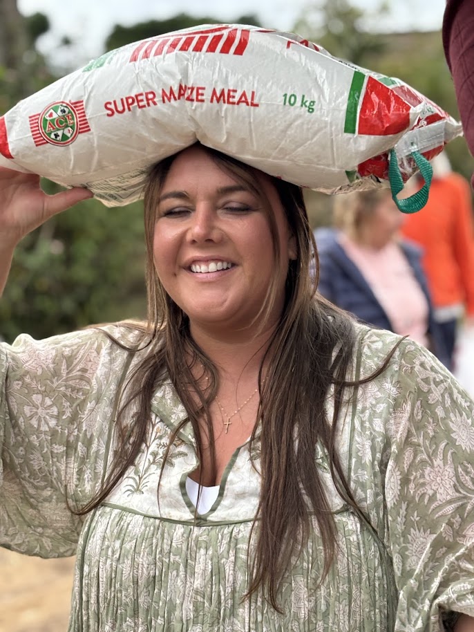 A long-time mission friend laughing as she balances a 10 kg maize meal bag on her head