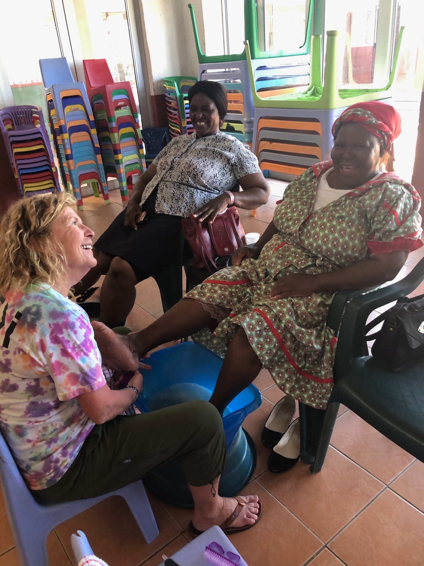 A visiting friend gently washing an elder woman's feet while two women from the community sit nearby smiling together