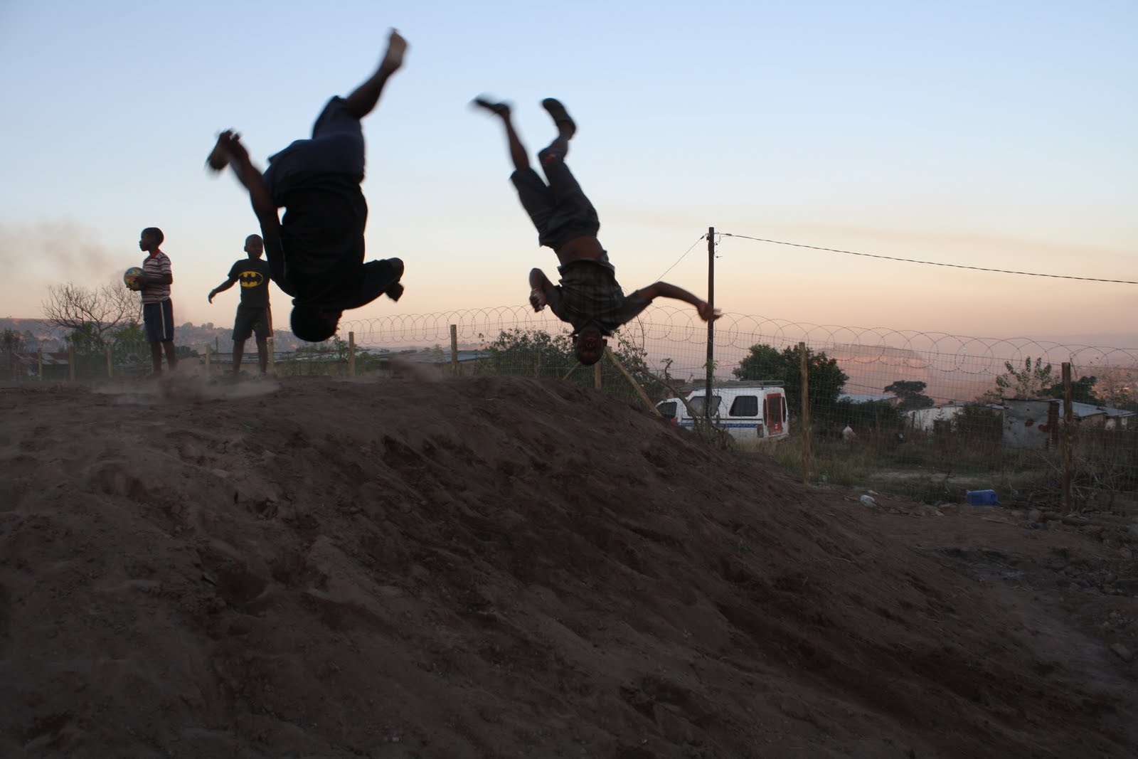 Two Ntshongweni boys mid-backflip off a red dirt mound at sunset with friends watching