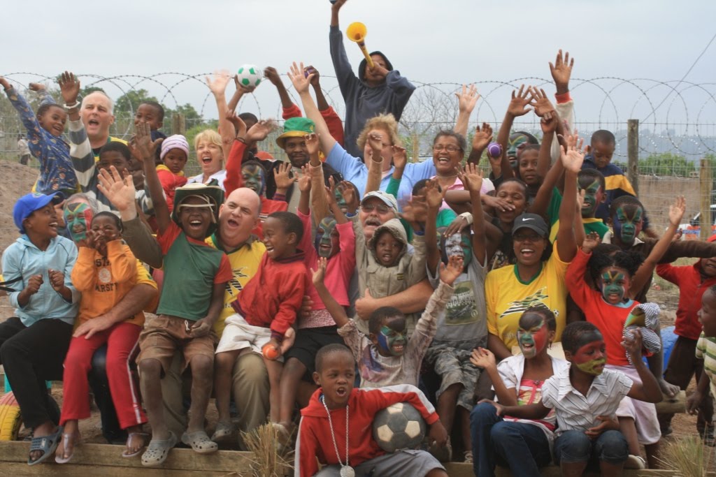 The first visiting group cheering together with face-painted Ntshongweni children behind a fence at sunset